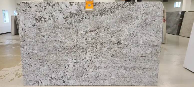This image shows a slab of Alaska White granite with a polished finish. The slab has a light color with subtle gray, brown, and black specks. Alaska White granite is a popular choice for countertops, backsplashes, and other interior applications. It is known for its beauty, durability, and low maintenance.